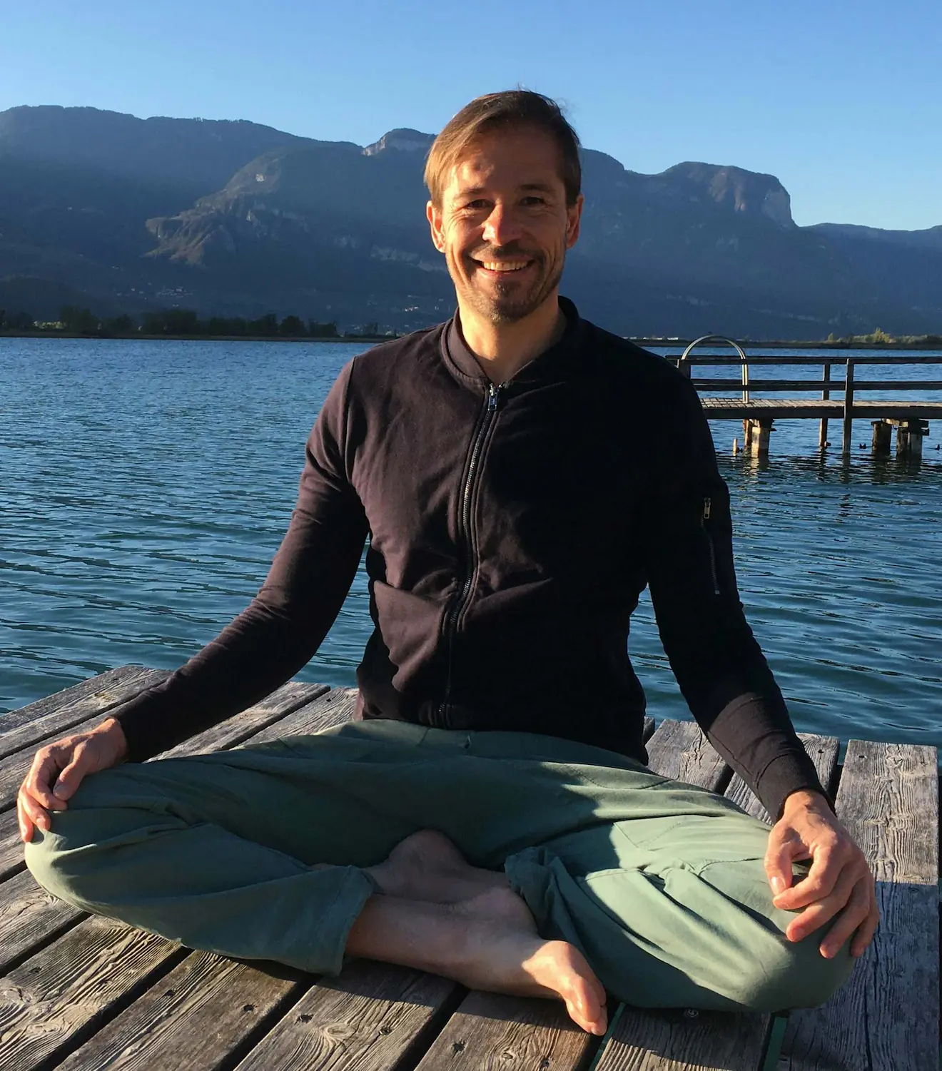 Yoga Vorarlberg Trainer Christoph meditiert auf einer Holzplattform neben einem malerischen See.